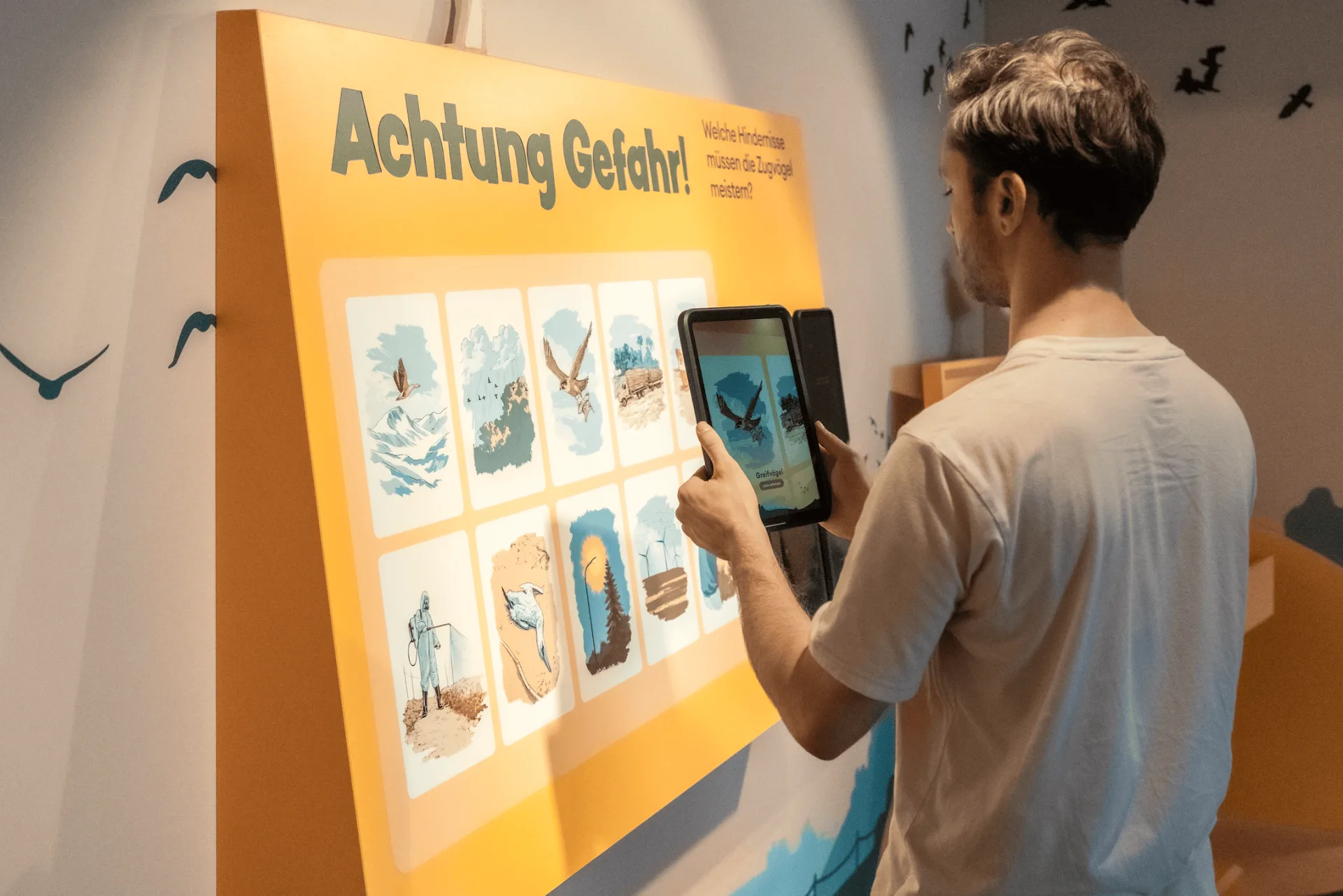 Eine Person hält ein Tablet und richtet die Kamera auf die Ausstellungsstation, um die dort platzierten Illustrationen zu scannen.