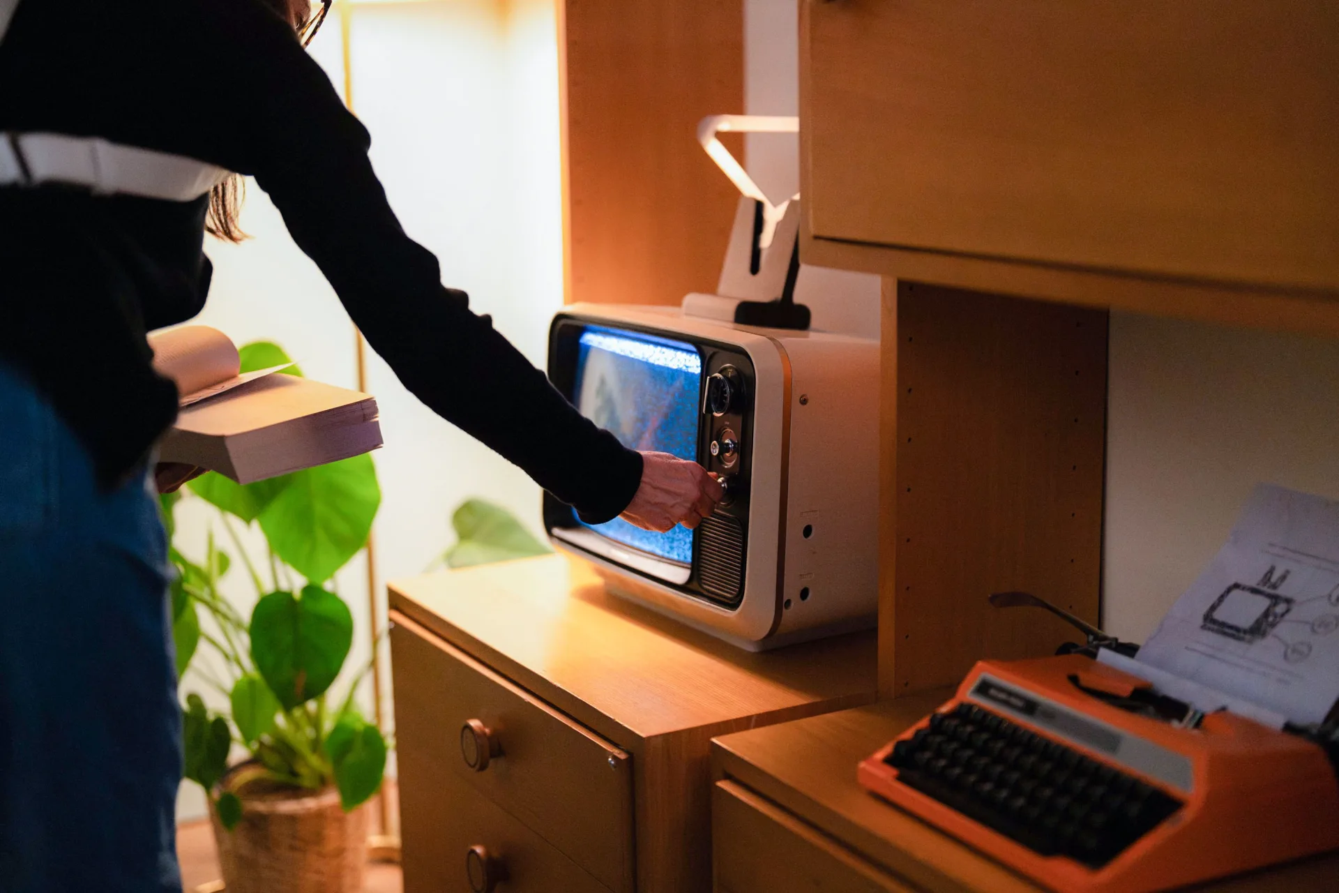 Person dreht im abgedunkelten Wohnzimmer-Set an einem Röhrenfernseher mit Bildrauschen; oben steckt ein Kleiderbügel als «Antenne», daneben eine Schreibmaschine.
