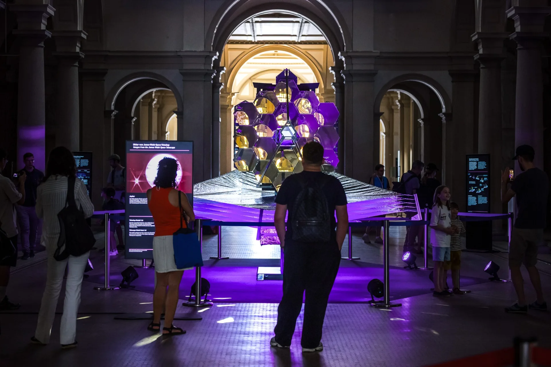 Frontale Nahaufnahme der goldenen Hexagonalspiegel des Teleskopmodells. Besucher:innen stehen davor und betrachten die Ausstellungsstücke und eine digitale Infostele auf der linken Seite.