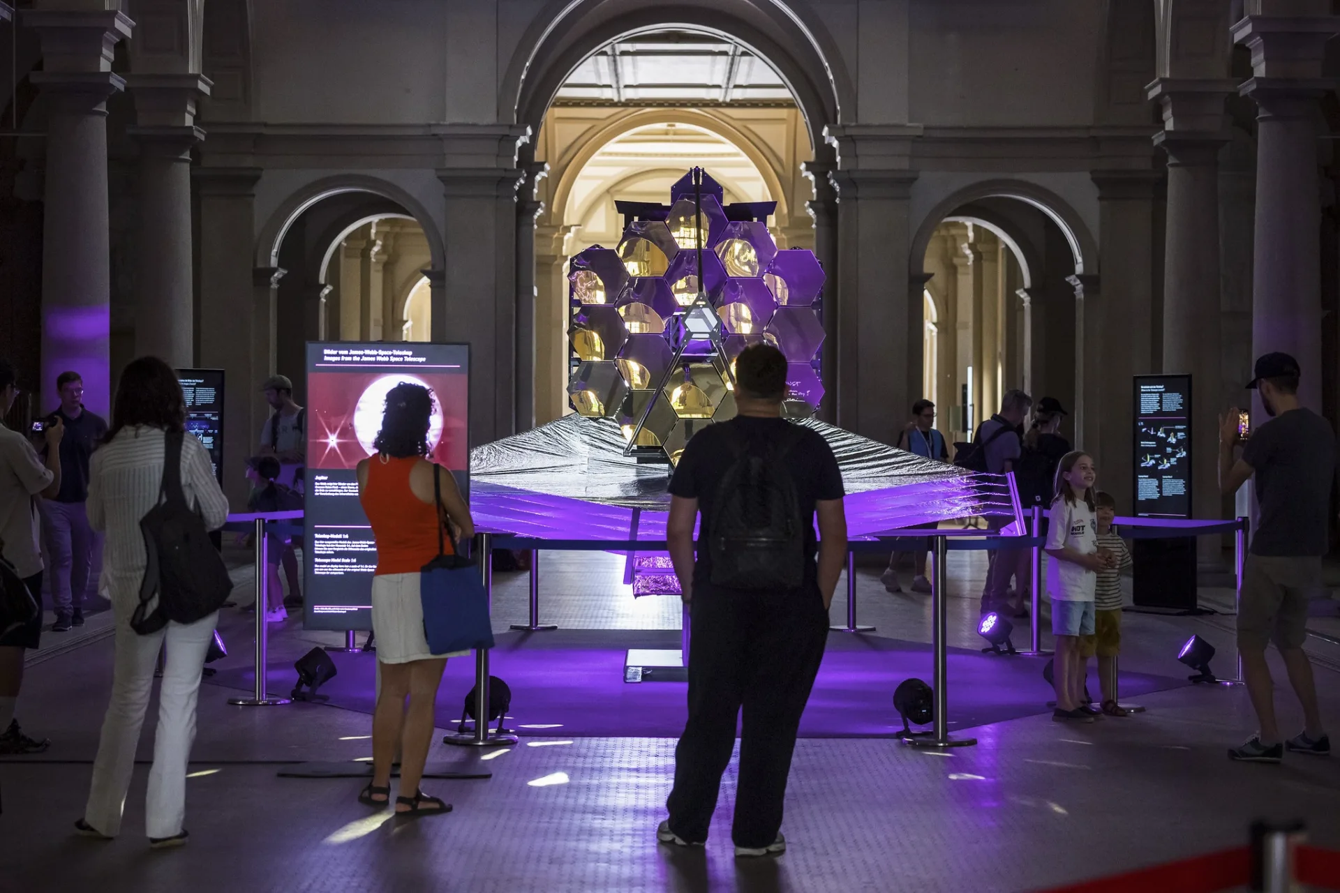Frontale Nahaufnahme der goldenen Hexagonalspiegel des Teleskopmodells. Besucher:innen stehen davor und betrachten die Ausstellungsstücke und eine digitale Infostele auf der linken Seite.