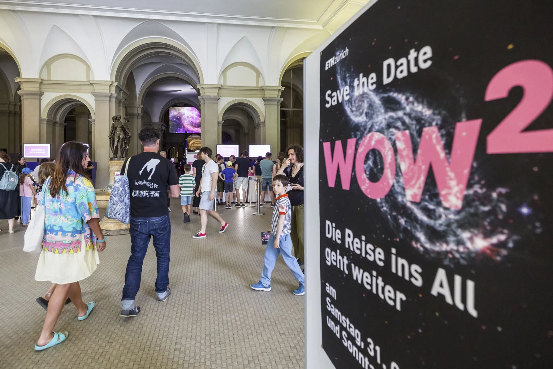 Eingangsbereich zum ETH-Event mit einem Plakat im Vordergrund, das die Aufschrift «WOW² – Die Reise ins All geht weiter» trägt, während Besucher:innen in die Ausstellungshalle gehen.