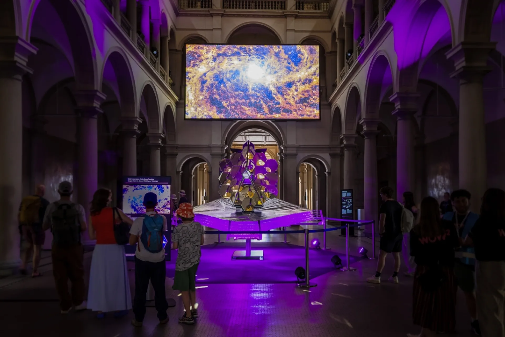 Symmetrische Gesamtansicht der Inszenierung in der Haupthalle. Das Teleskopmodell steht zentriert unter der grossen Leinwand mit Galaxie-Aufnahme, während Besucher:innen links den synchronisierten Informationsscreen studieren.