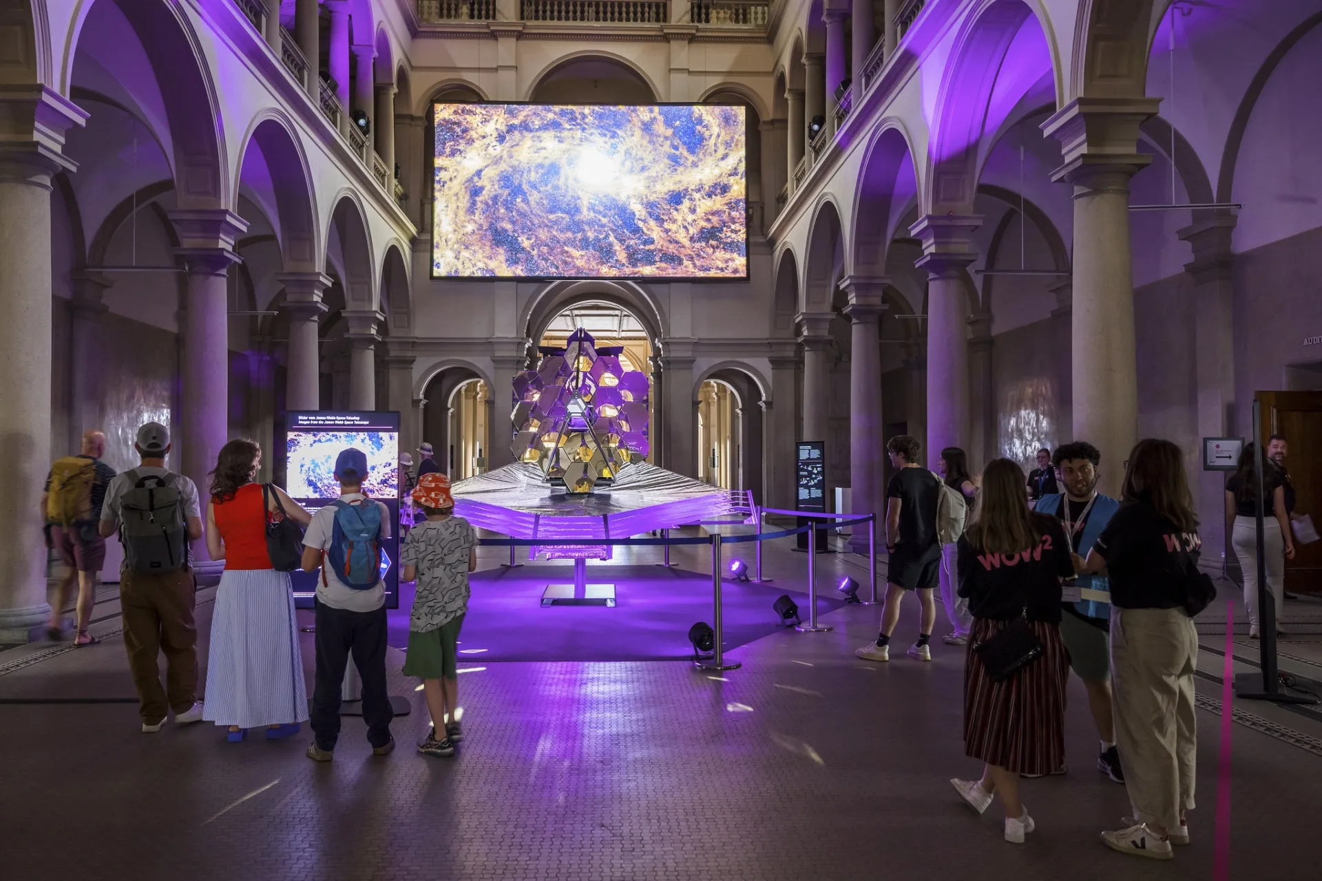 Symmetrische Gesamtansicht der Inszenierung in der Haupthalle. Das Teleskopmodell steht zentriert unter der grossen Leinwand mit Galaxie-Aufnahme, während Besucher:innen links den synchronisierten Informationsscreen studieren.