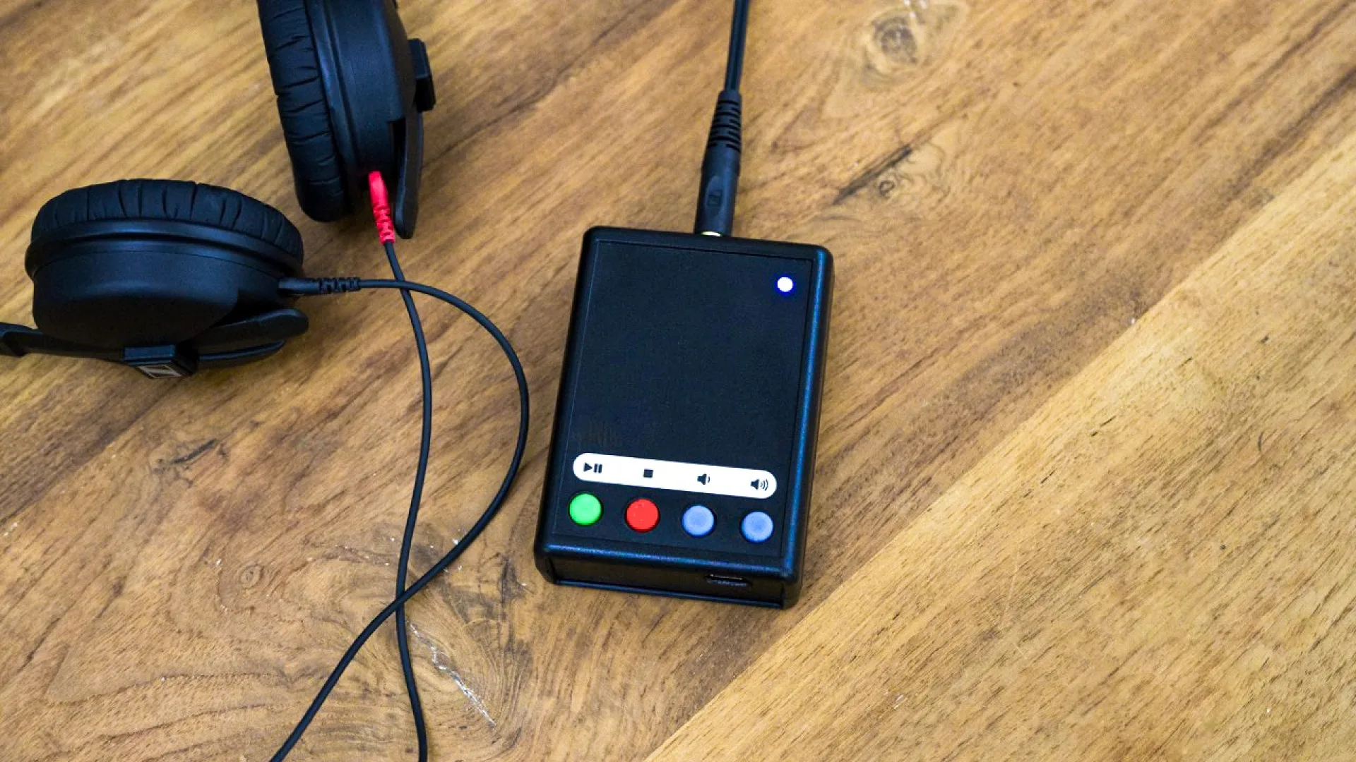 Lucid Audioplayer. Ein schwarzes Kunststoffgehäuse mit vier farbigen Knöpfen liegt auf einem Holztisch. Ein Kopfhörer mit Kabel liegt daneben.